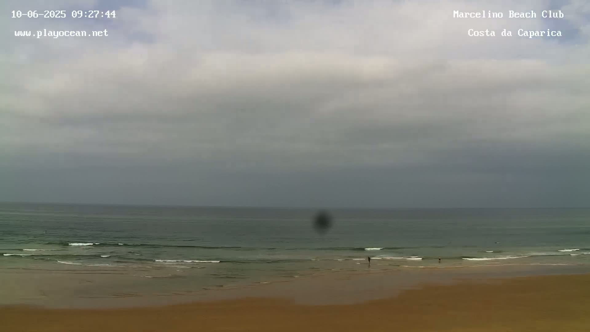 A mostly cloudy day at a beach shows a calm ocean with small waves lapping a wide, sandy shore where a few people are visible.