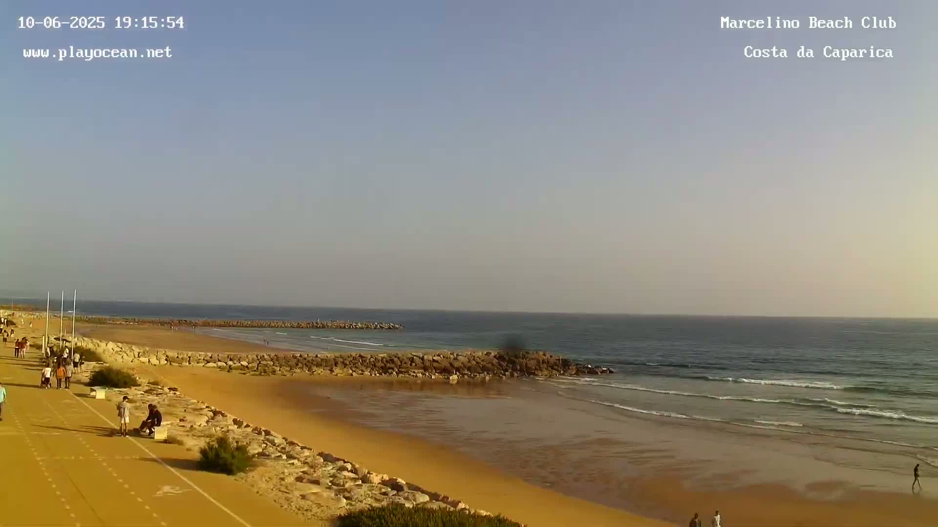 A sandy beach with calm waves and a few people walking along the shore under a clear, light-blue sky.