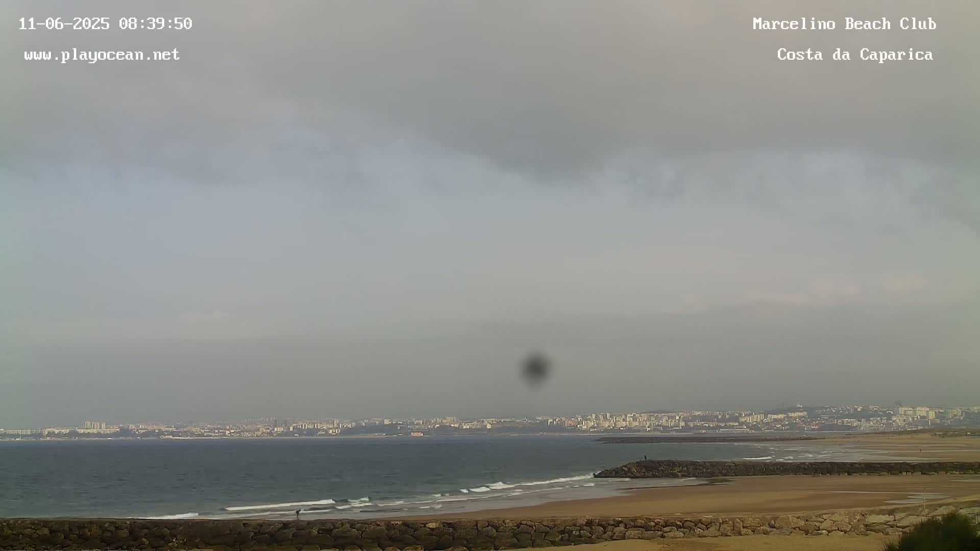 A hazy overcast sky hangs over a calm ocean beach with a distant cityscape visible on the horizon.