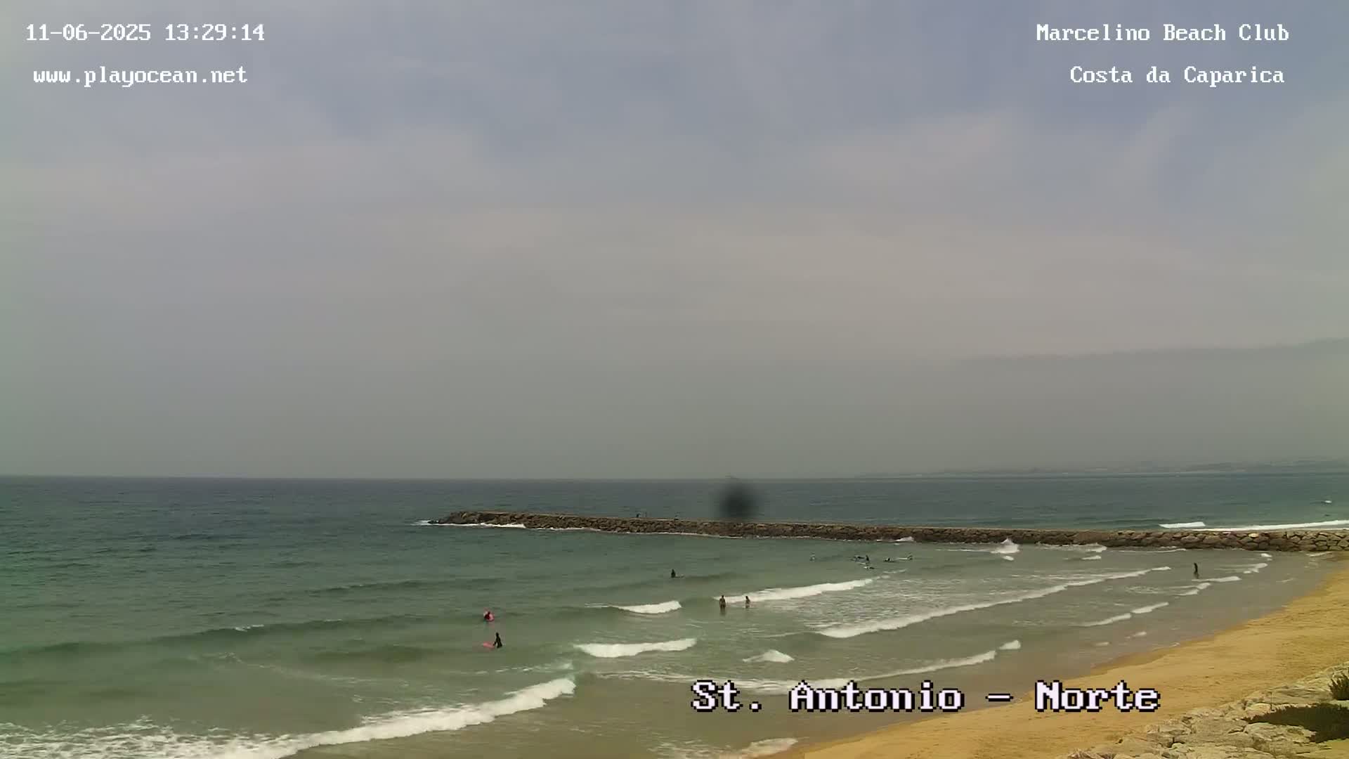 A beach scene shows a calm ocean with a few surfers and a rock jetty under an overcast sky.