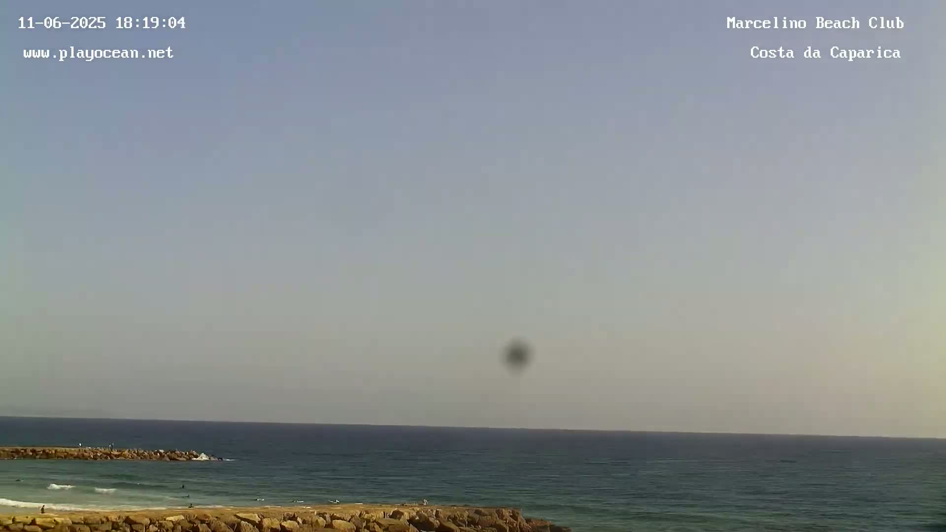 A mostly clear sky overlooks a calm ocean with a rocky shore and a few people in the water.