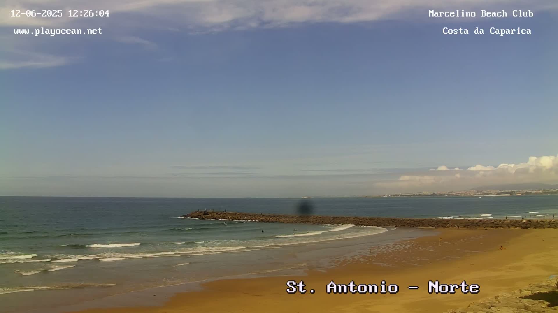 A sandy beach with small waves and a rocky breakwater stretches along a calm ocean under a mostly clear, sunny sky.