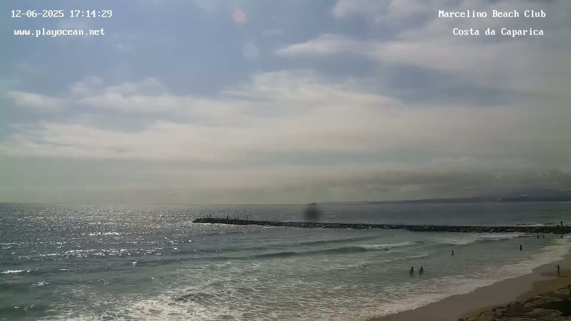 A partly cloudy day at a beach shows a long jetty extending into a wavy ocean, with a few people swimming and walking along the sandy shore.