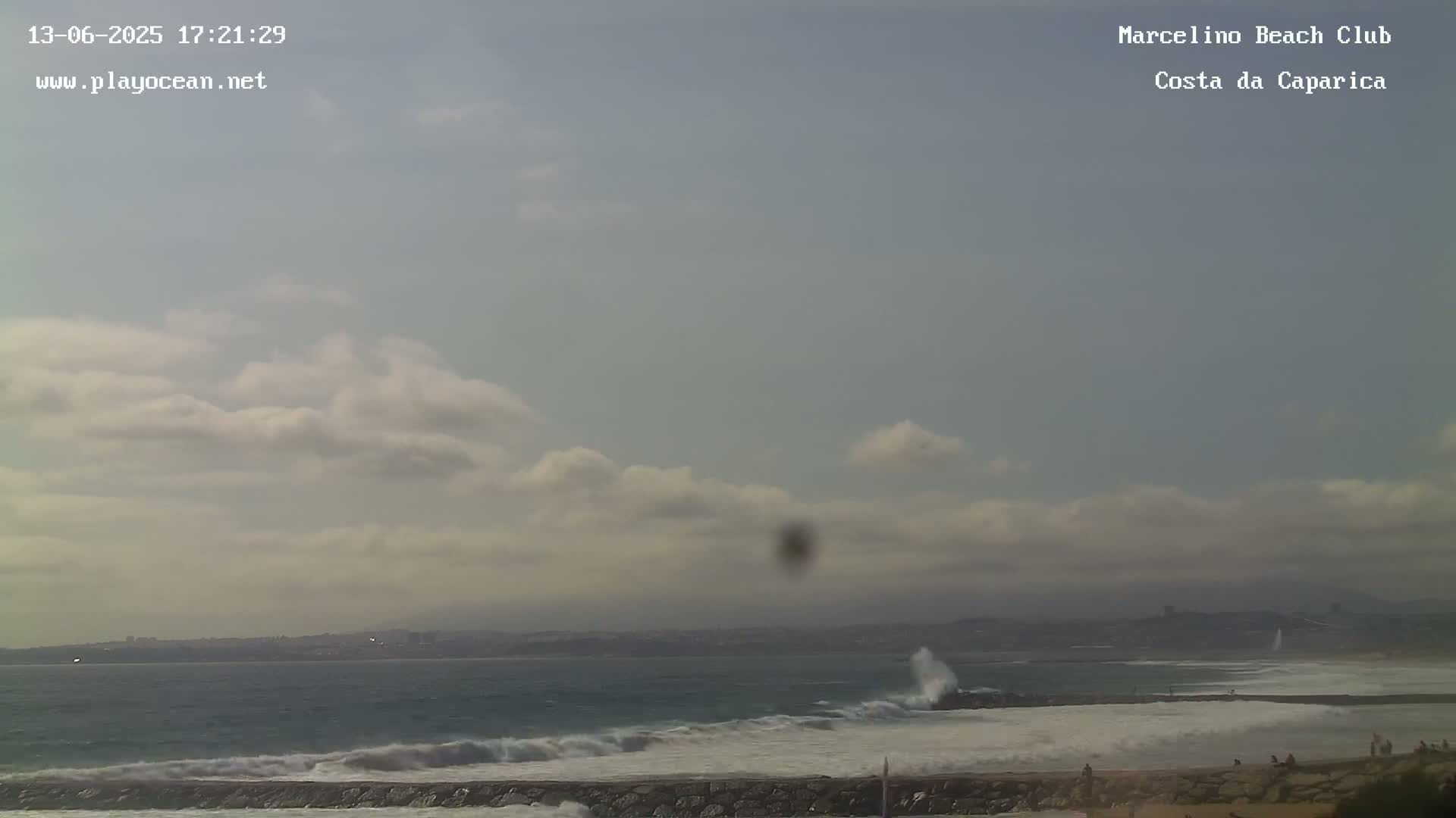 A partly cloudy sky overlooks a rocky coastline with waves crashing against a breakwater, and a few people visible on the beach.