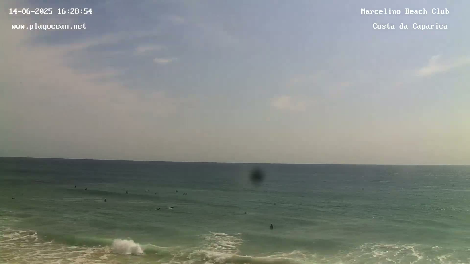 A mostly clear, slightly hazy sky overlooks a calm ocean with several surfers visible in the water, small waves lapping the shore.