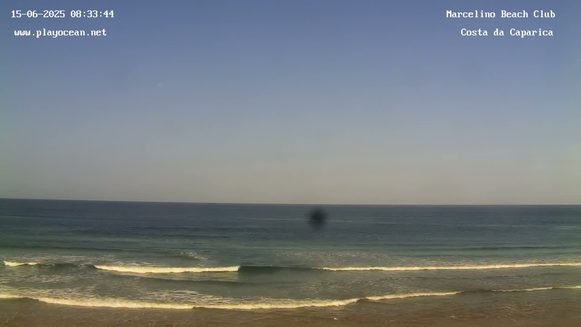 A mostly clear sky overlooks a calm ocean with small waves breaking on a sandy beach.