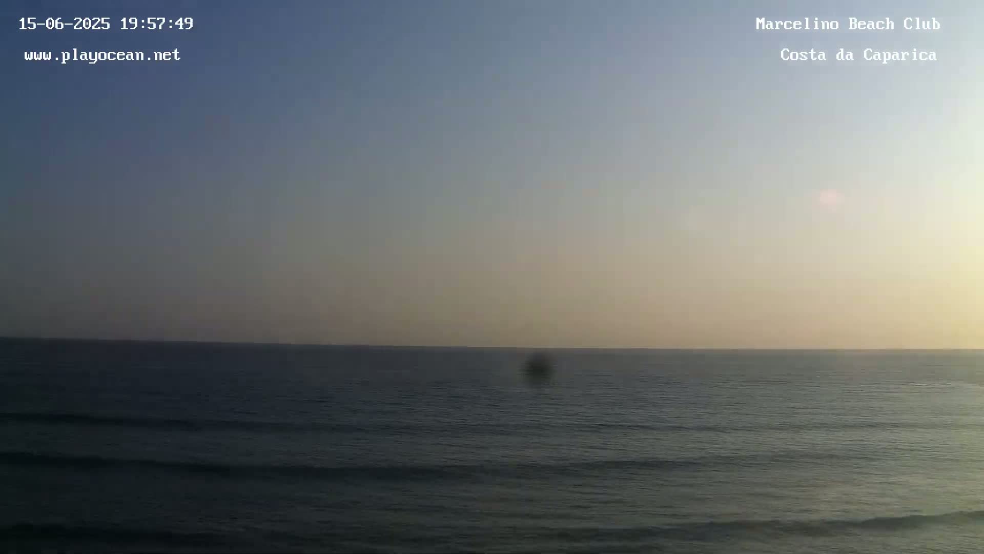 A hazy, sunny day at a beach shows a calm ocean with small waves lapping a sandy shore where people are swimming and sunbathing, with a city visible in the distance.
