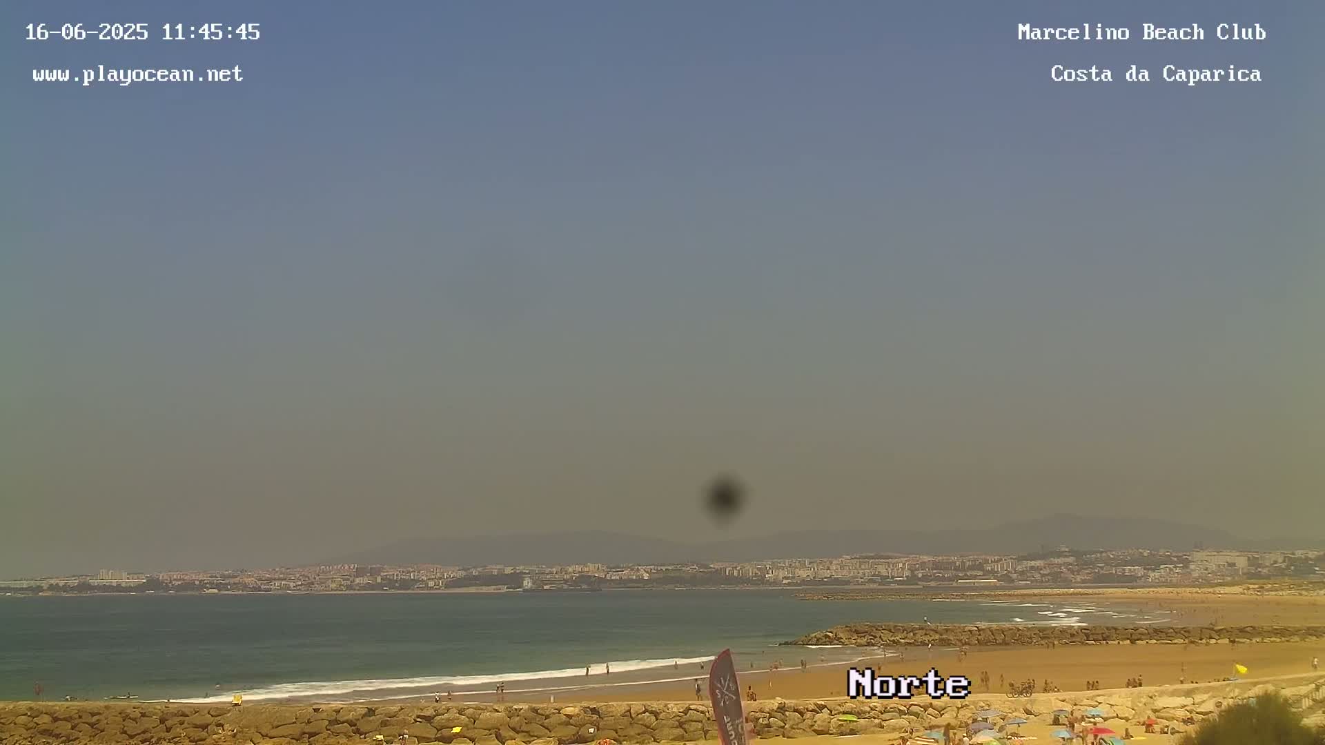 A hazy, sunny day at a beach shows a sandy shore with people, calm waves lapping the sand, and a distant cityscape under a pale blue sky.