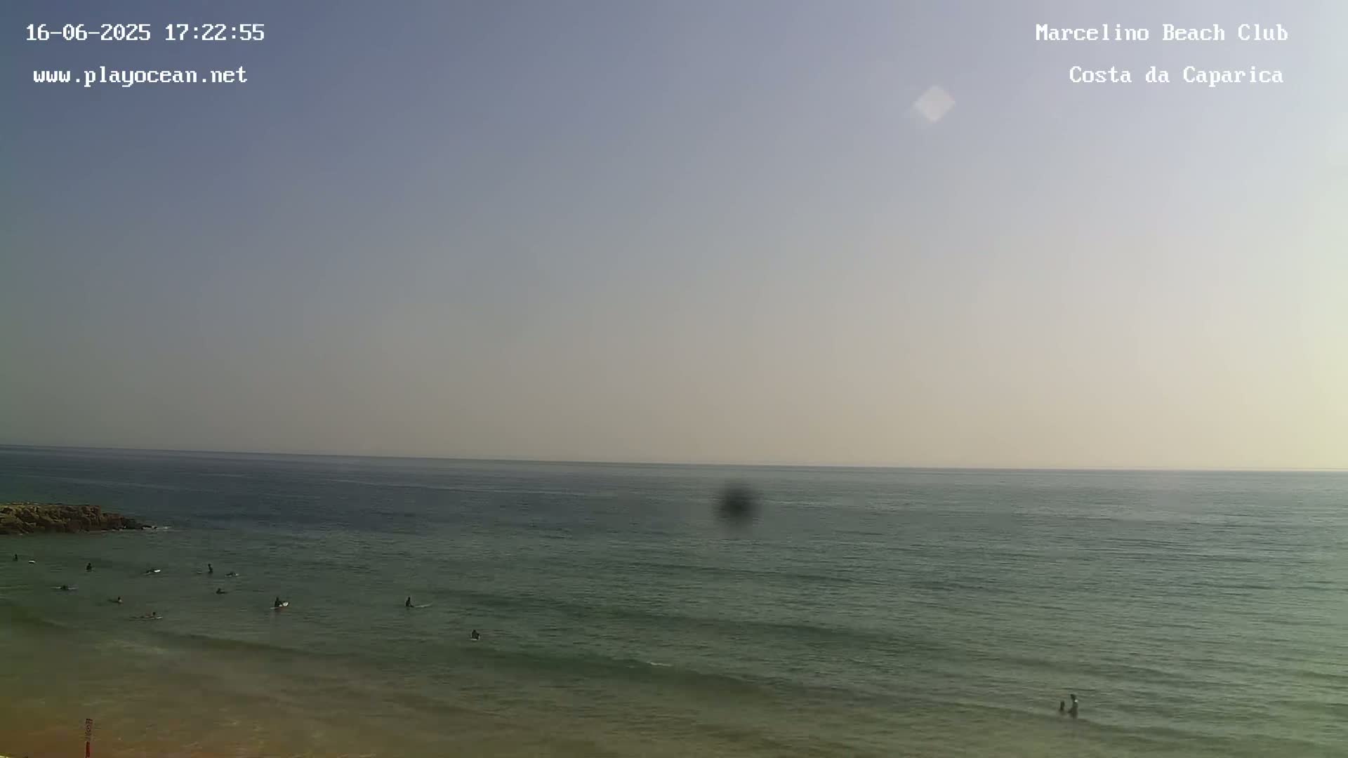 A mostly clear, hazy sky overlooks a calm ocean with several surfers in the water near a sandy beach.