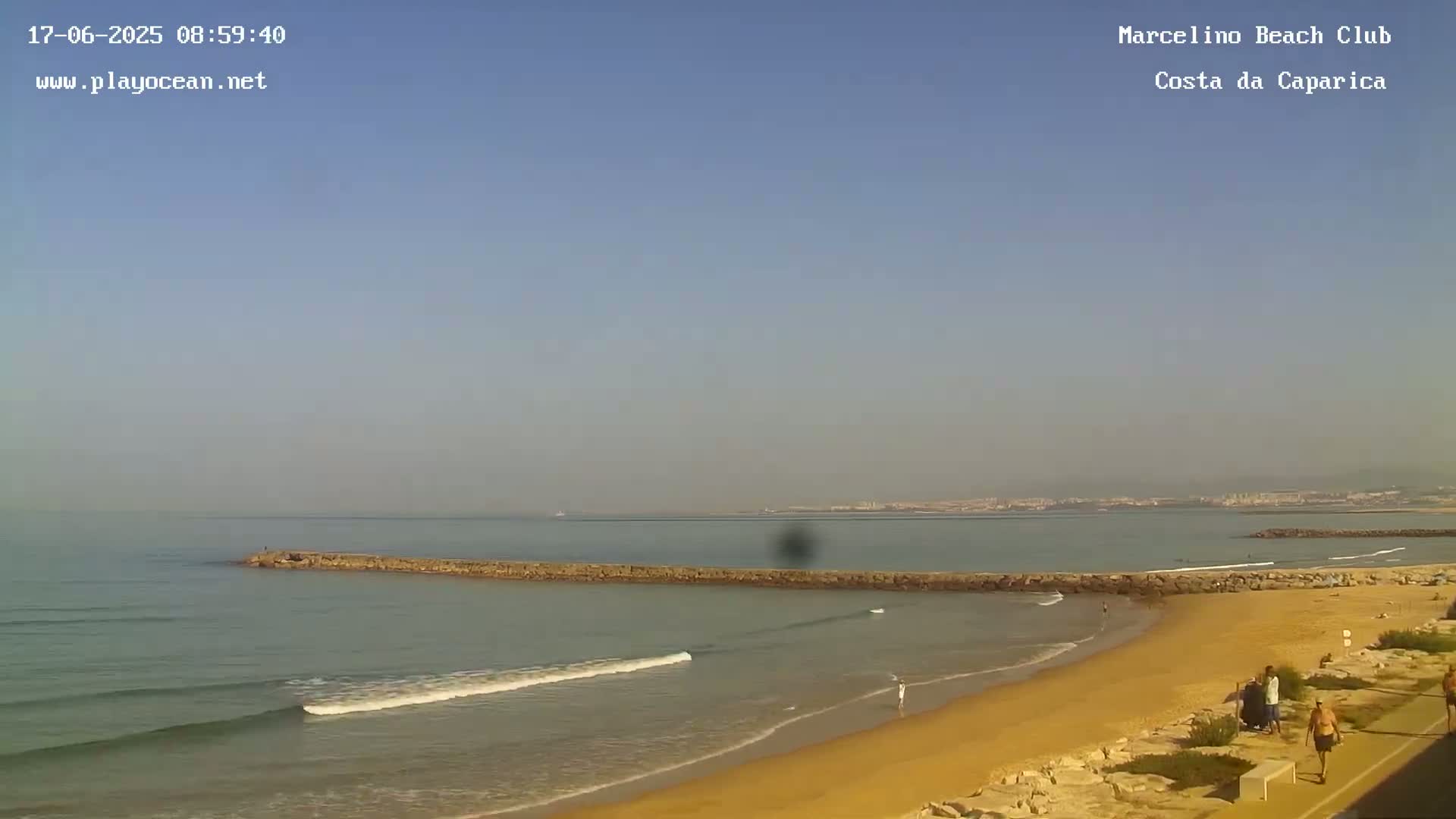 A sandy beach with a few people on it is separated from calmer ocean water by a long, low rock wall under a clear, light blue sky.