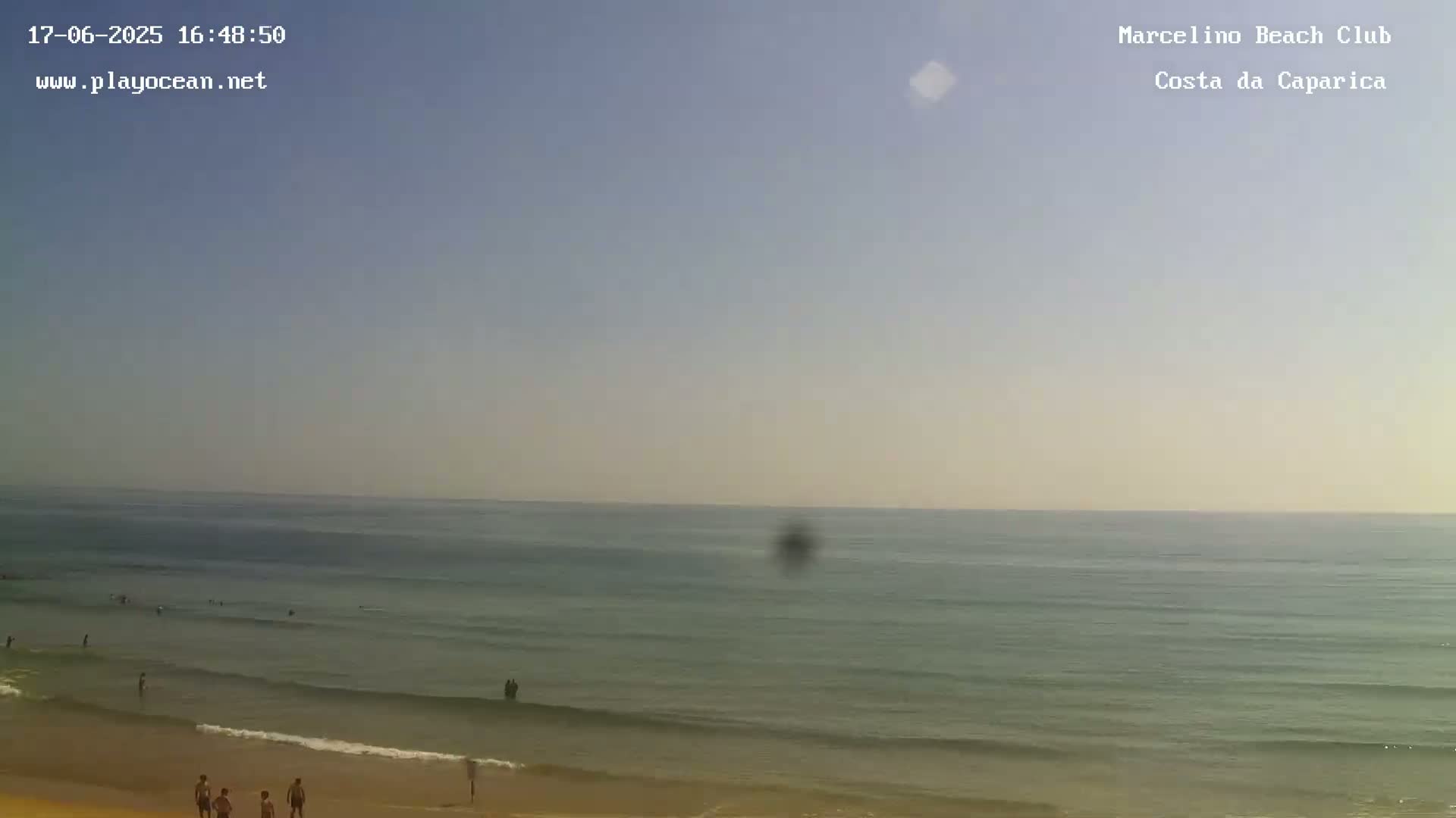 A mostly clear, sunny day at a beach with calm, light-blue water and a few people on the sand.