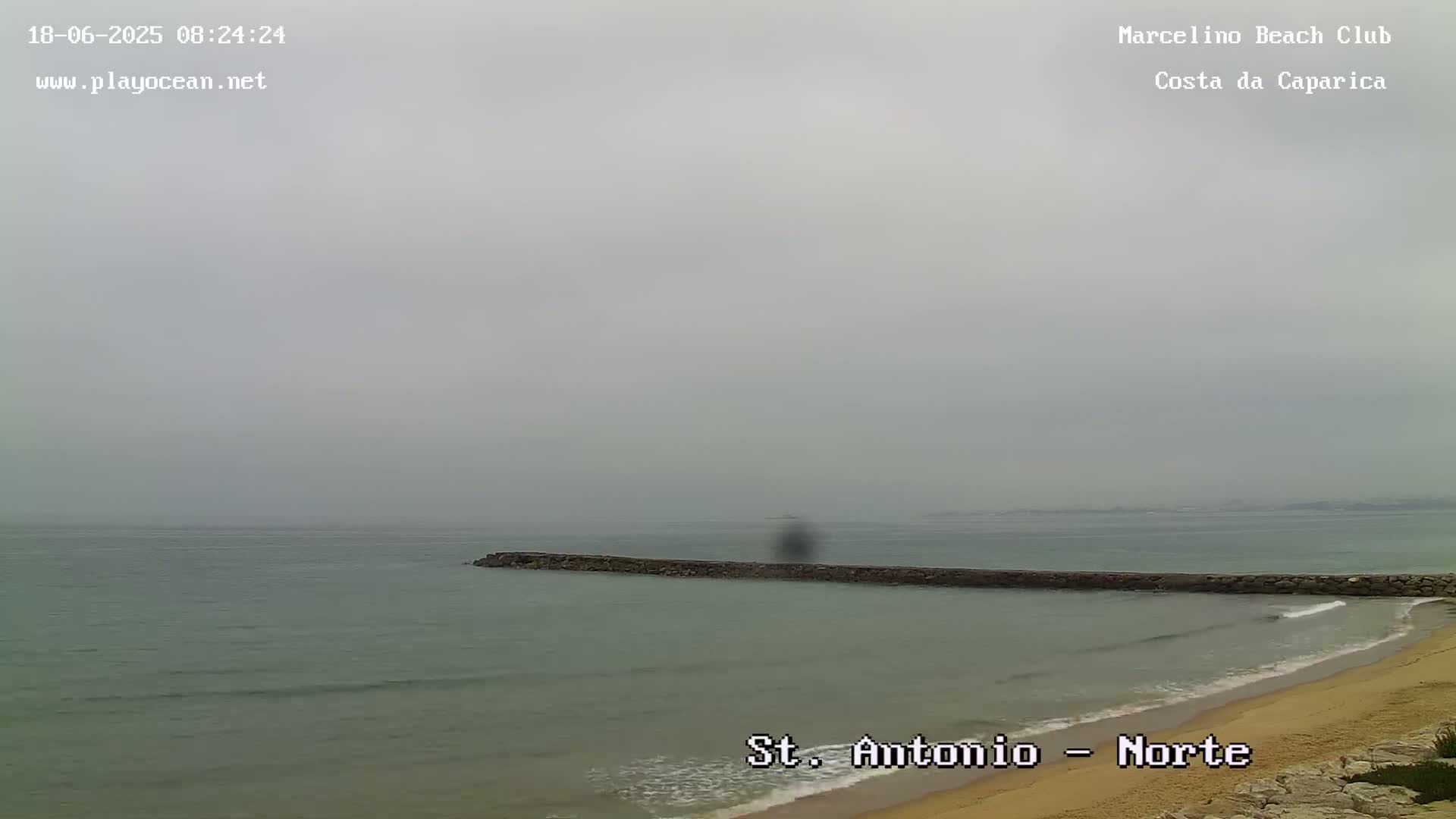 A foggy, overcast day reveals a long, dark breakwater extending into a calm, gray-green ocean next to a sandy beach.