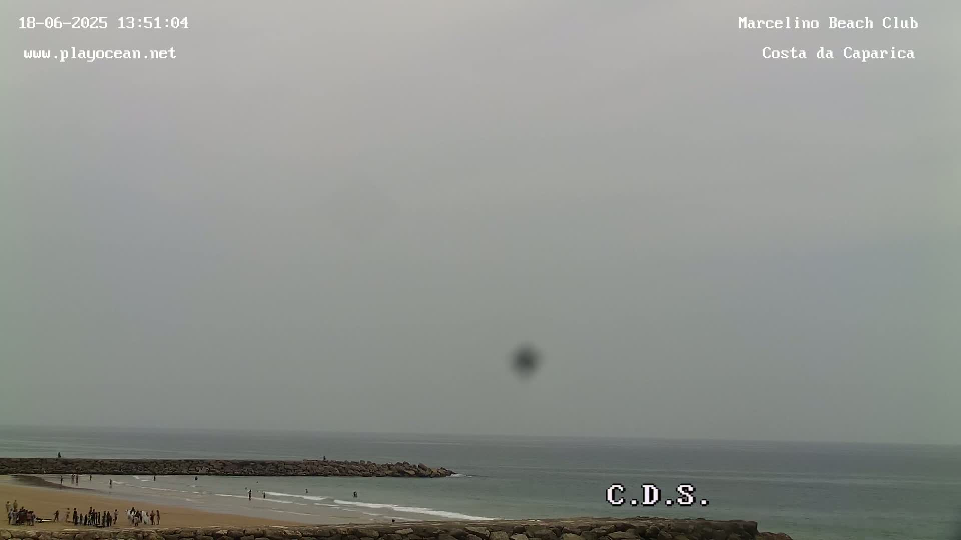 A hazy overcast sky hangs over a beach scene with a few people on the sand and in the water, a rock wall separating the beach from the ocean.