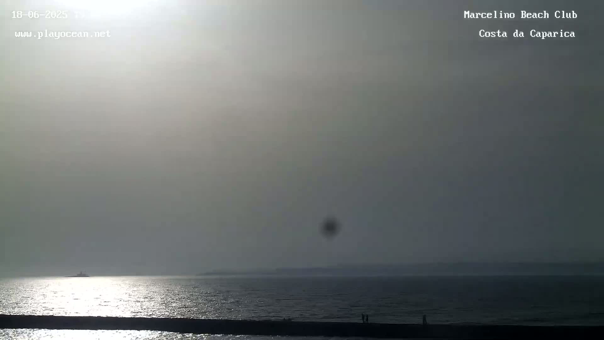 A hazy, bright day shows a calm ocean with a distant shoreline and a few figures on a dark jetty.