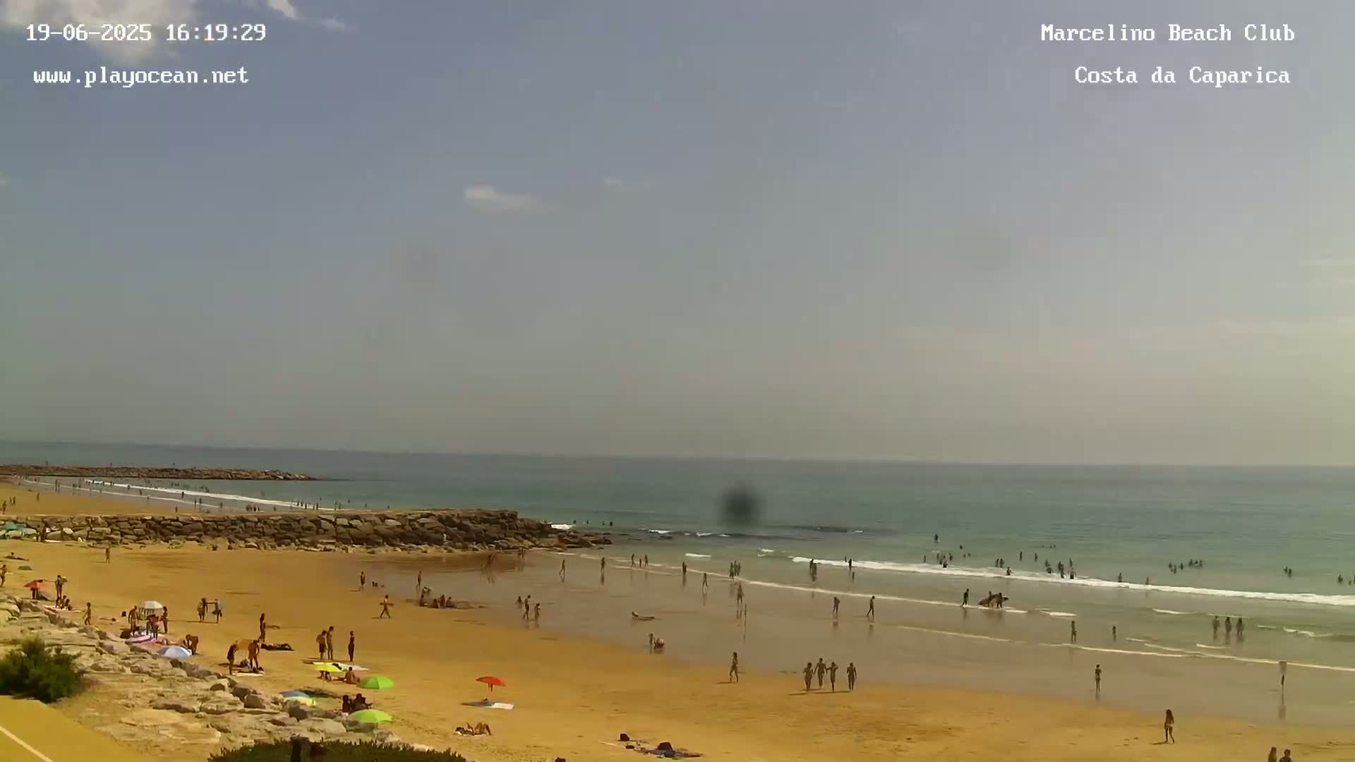 A sunny day at a sandy beach shows many people swimming and sunbathing near the ocean under a mostly clear sky.