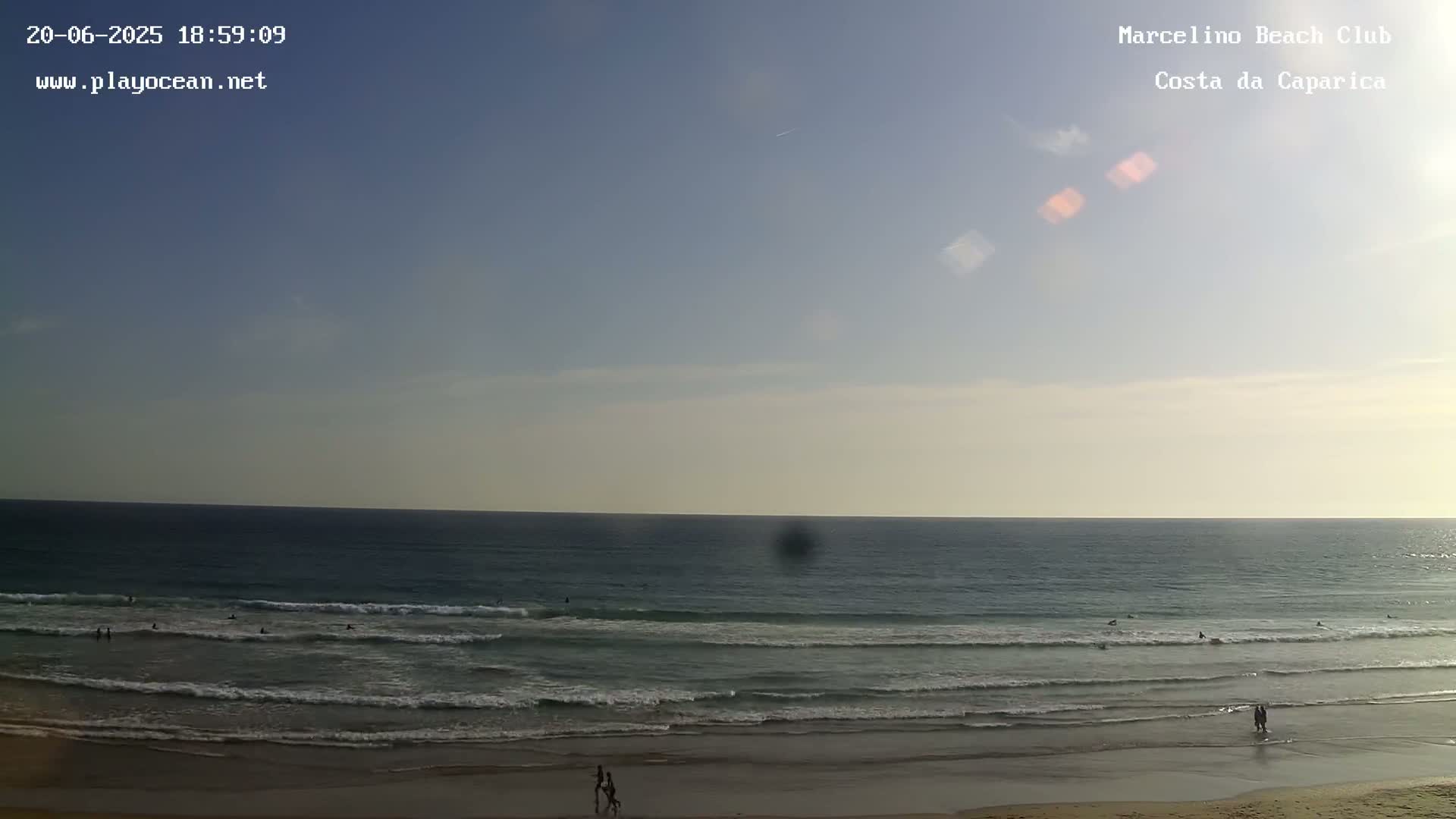 A mostly clear sky over a calm ocean with small waves breaking on a sandy beach, where several people are visible on the shore and in the water.