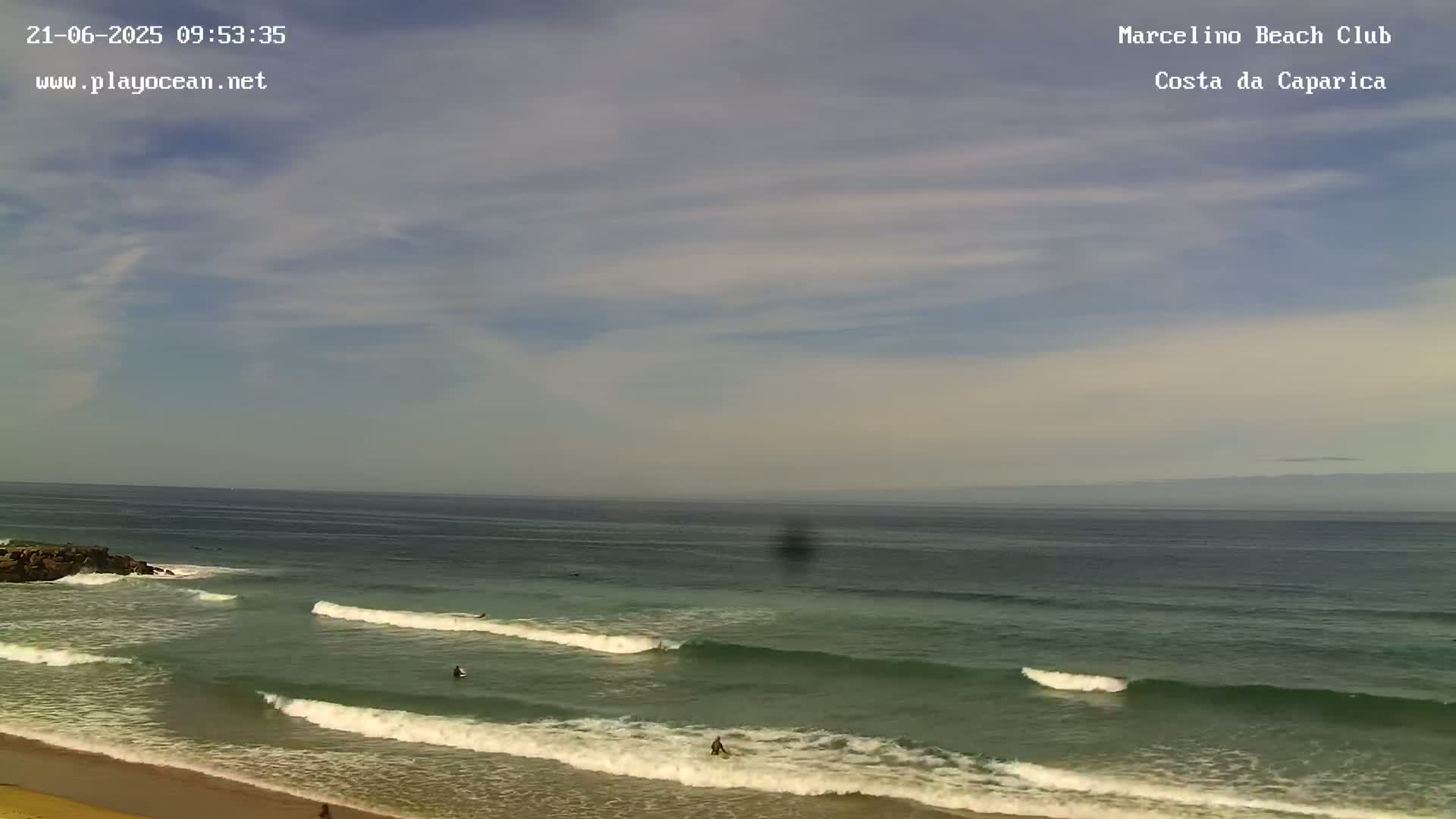 A mostly sunny day shows a sandy beach with small waves rolling onto the shore, where a few surfers are visible in the ocean.