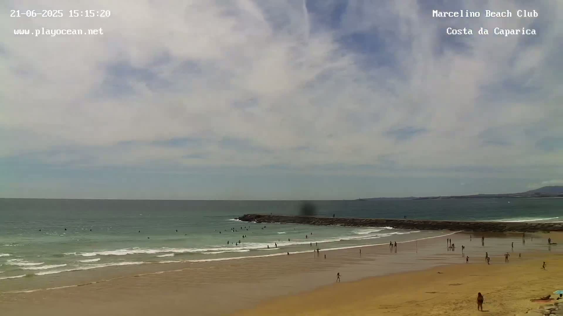 A sandy beach with a calm ocean and a few people swimming and sunbathing under a partly cloudy sky.
