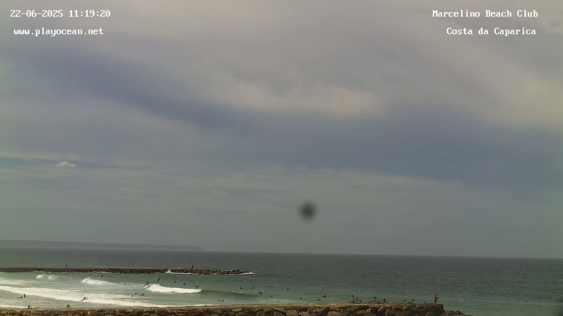 An overcast sky hangs over an ocean beach where several surfers are in the water near a rock jetty.