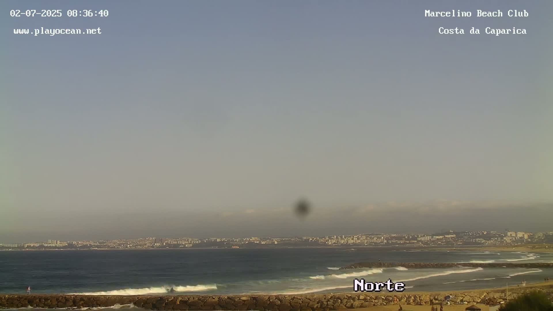 A hazy, overcast day reveals a calm ocean with small waves lapping a sandy beach, backed by a distant cityscape.