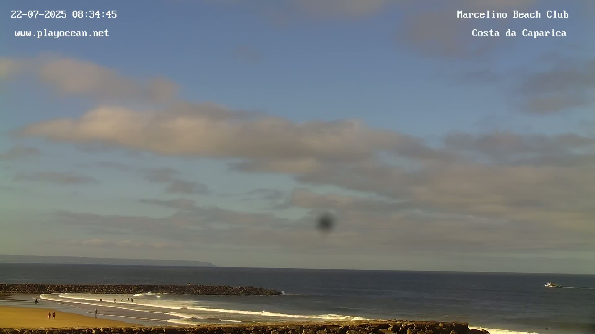 A mostly sunny day at a beach shows a calm ocean with small waves breaking near a rocky shoreline, and a few people are visible on the sandy beach.