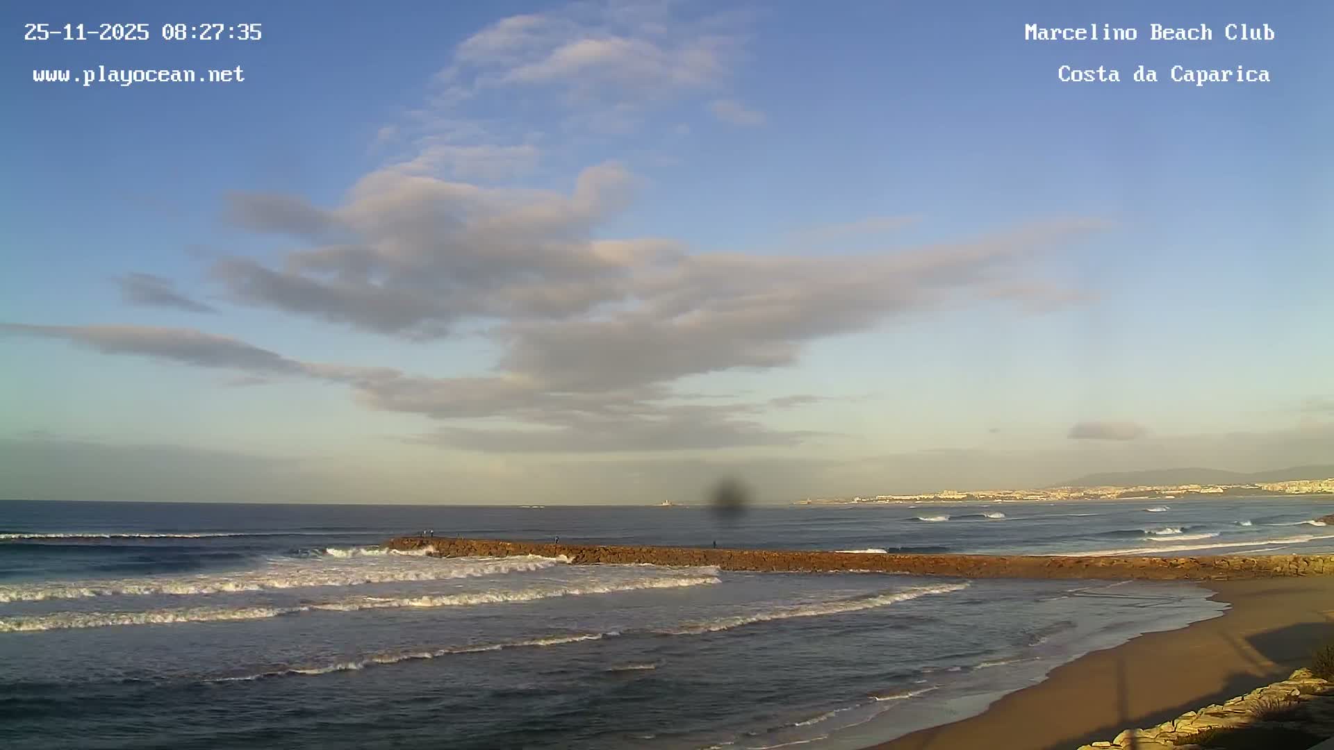 A partially cloudy sky hangs over a coastal scene with ocean waves breaking on a sandy beach and around a long rock jetty, with a distant city visible along the shore.