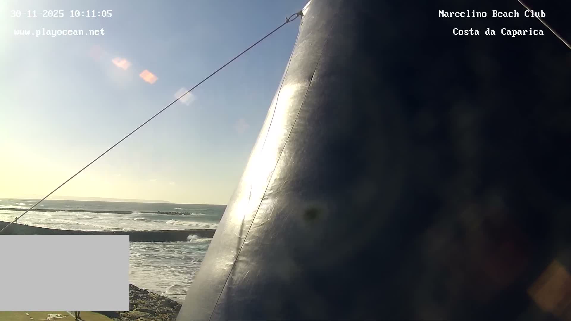 Under a bright, sunny sky, ocean waves crash against a breakwater with a distant coastline, as seen past a large, dark, curved surface dominating the right side of the frame.