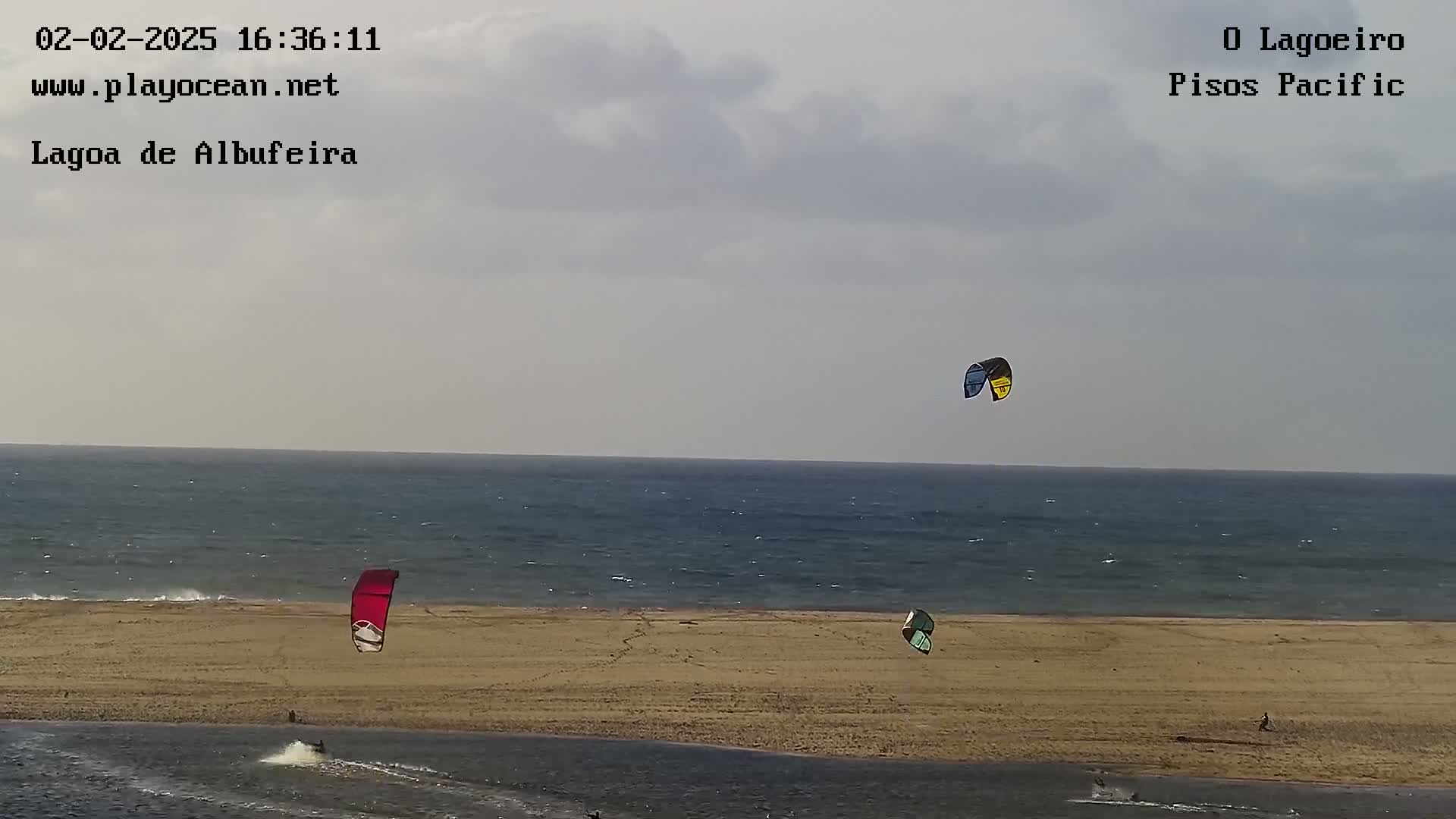 Three kitesurfers are visible on a sandy beach near a calm ocean under a mostly cloudy sky.