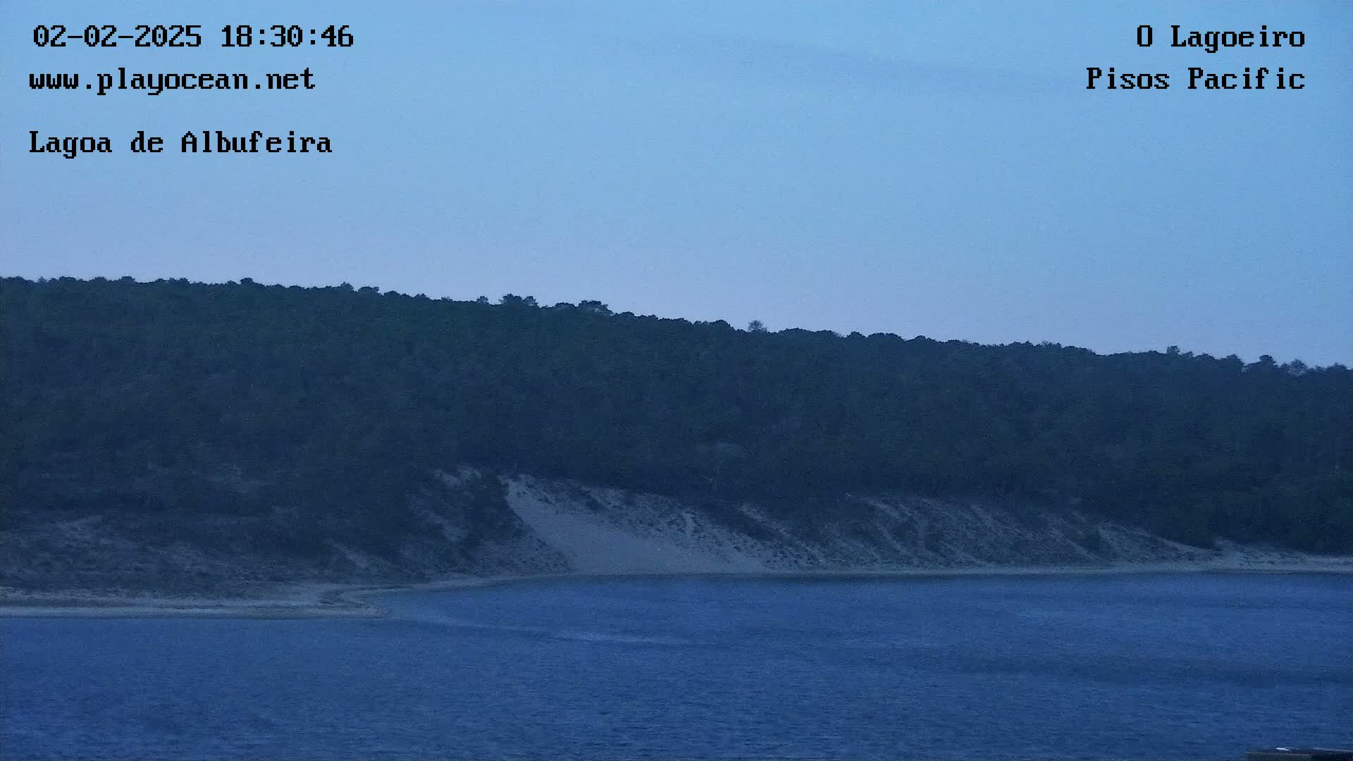 A dark, tree-covered hill slopes down to a sandy beach bordering a calm, blue body of water under a twilight sky.