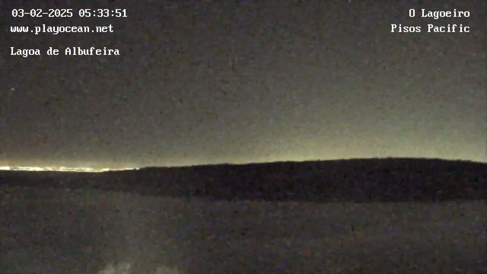 A nighttime, long shot shows a dark landscape with a distant horizon illuminated by city lights under a mostly clear, dark sky.