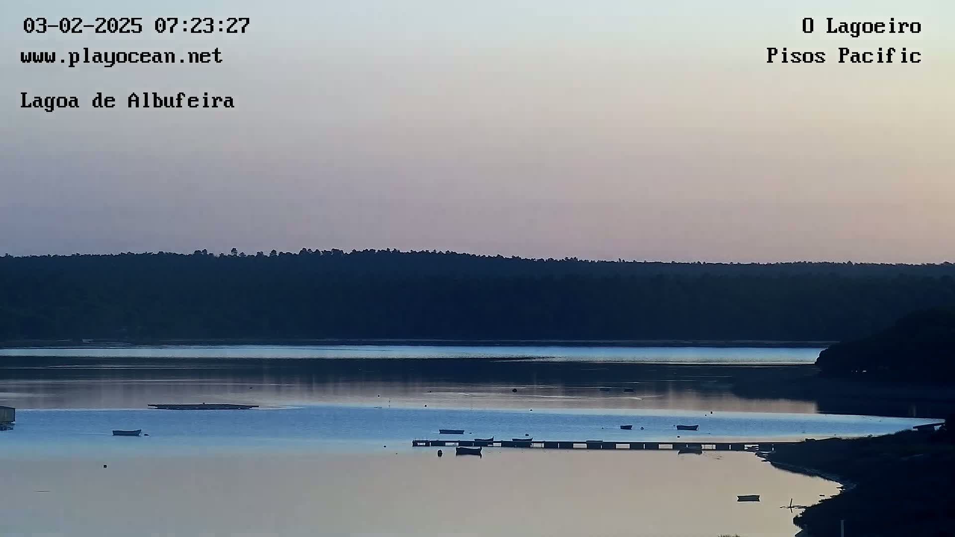 Several small boats are moored at a calm, twilight lagoon, with a dark treeline in the background under a pale, dusky sky.