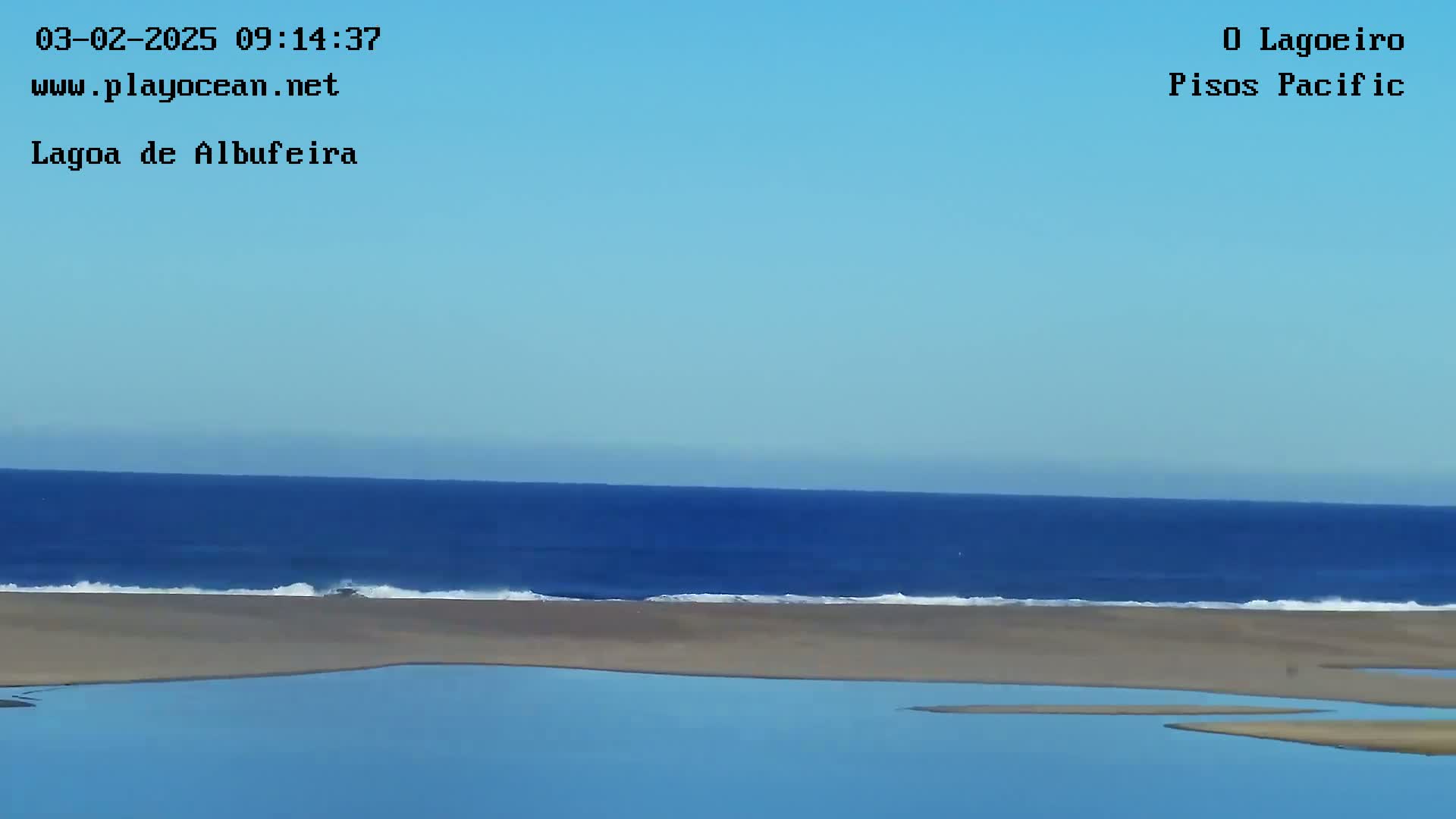 A calm ocean with a sandy beach and calm blue water in the foreground under a clear blue sky.