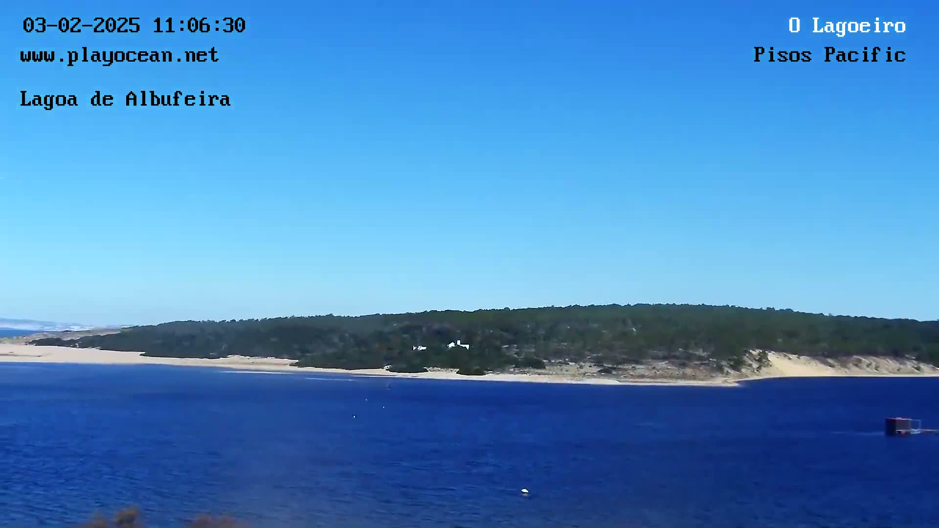 A tranquil body of blue water stretches before a sandy shore backed by a verdant, tree-covered hill under a clear blue sky.