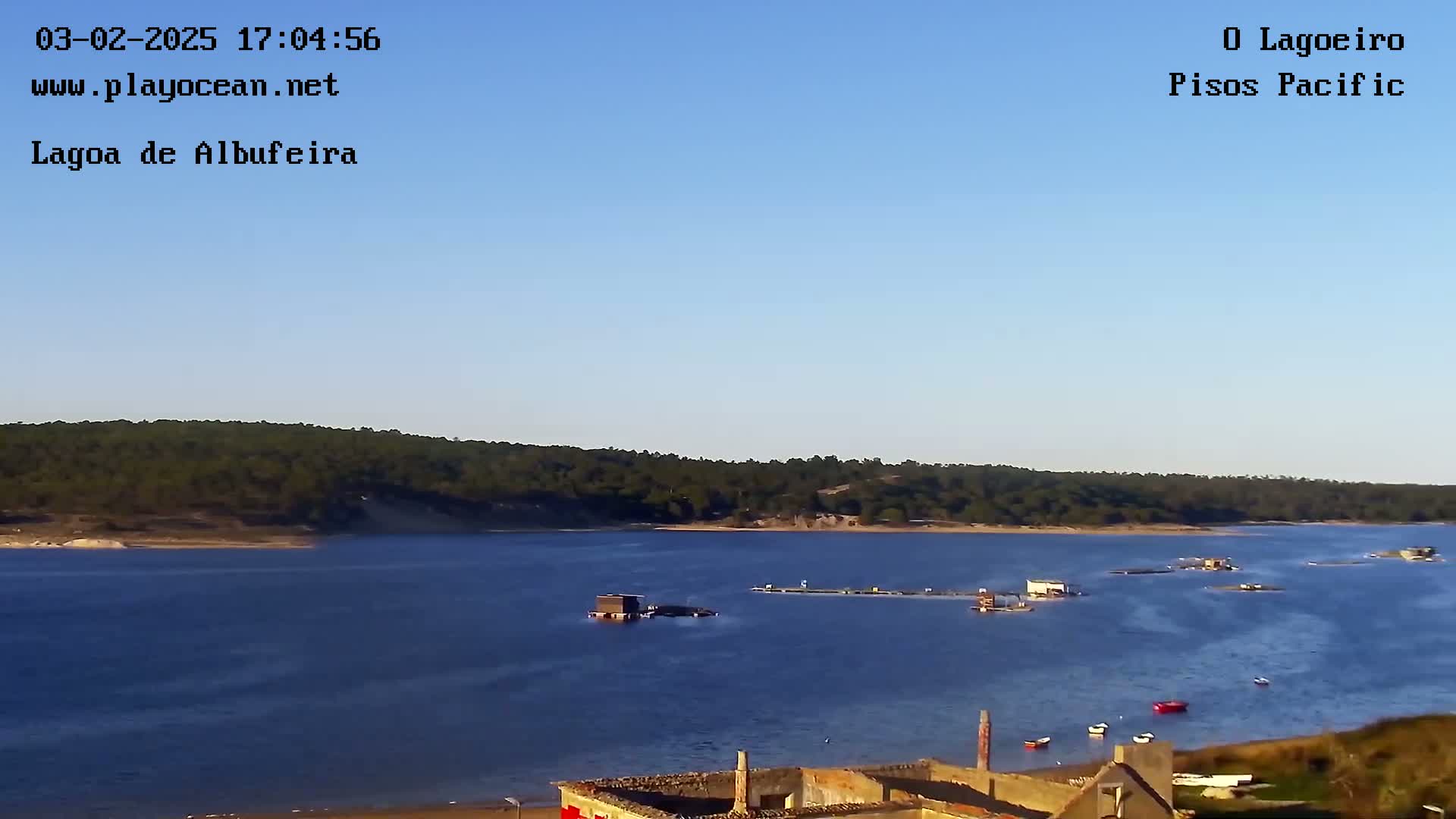 A calm body of water stretches out under a clear blue sky, with a few small structures and boats visible on the water's surface, and a line of trees and hills in the distance.