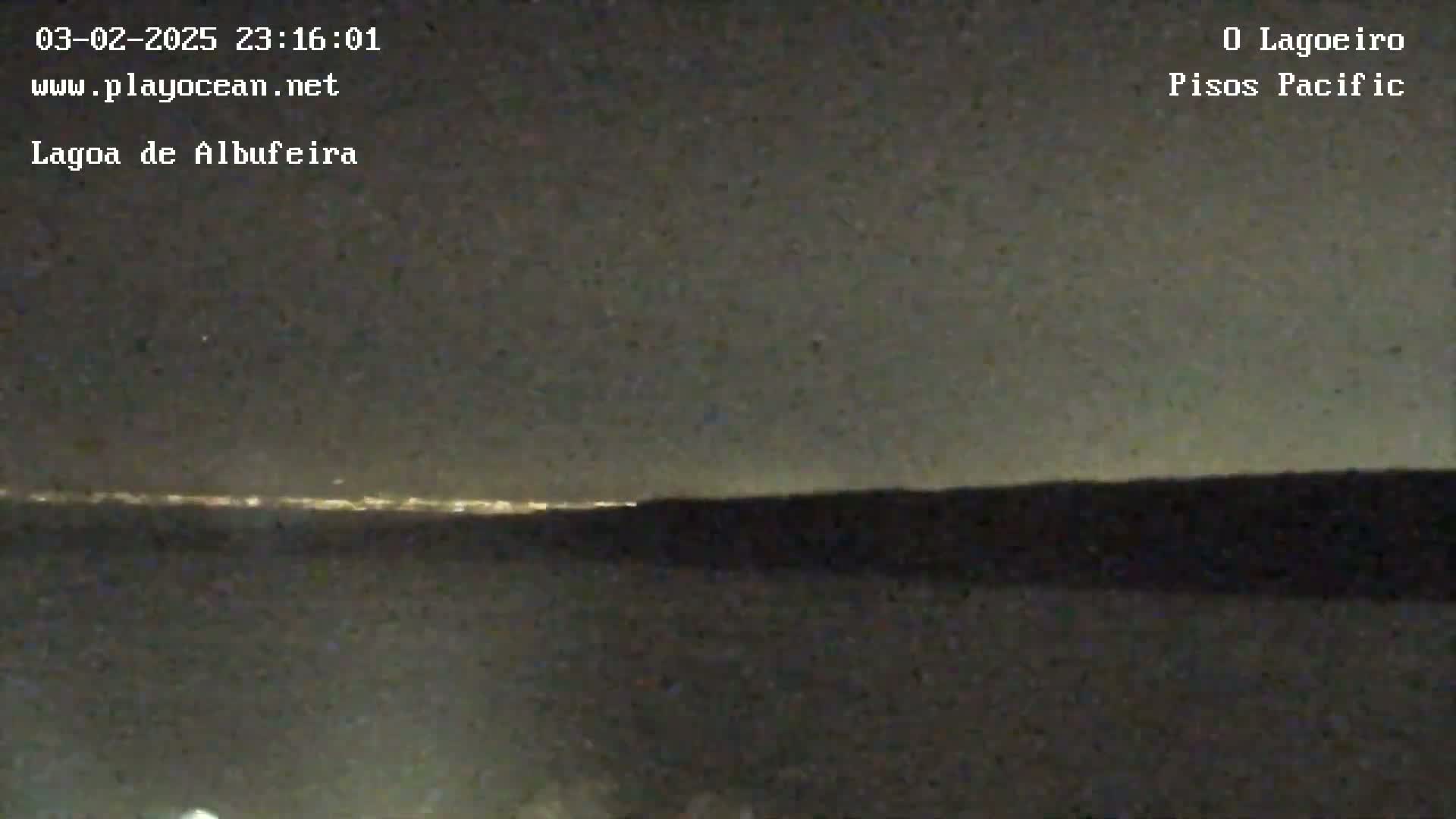 A nighttime long shot shows a dark landmass silhouetted against a dimly lit horizon with a faint glow of distant city lights under a mostly clear, dark sky.