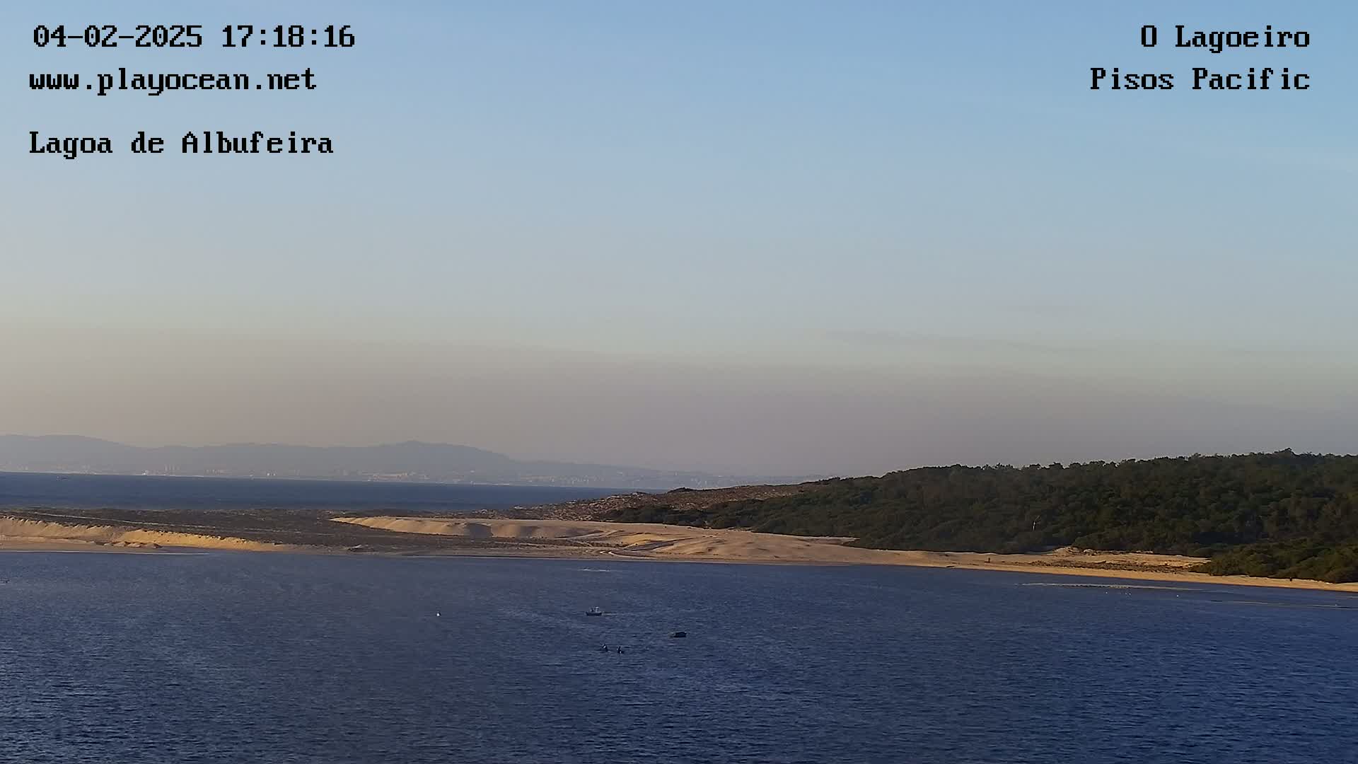 A calm body of water with sand dunes and a forested hillside on the far shore under a clear, light-blue sky.