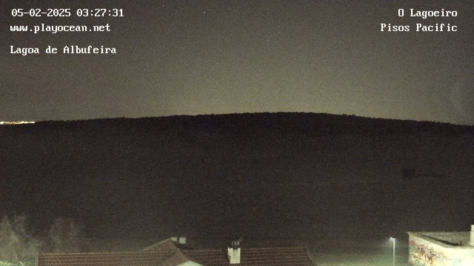 A dark, nighttime view shows a hill silhouetted against a dusky sky, with a few lights visible in the distance and rooftops in the foreground.