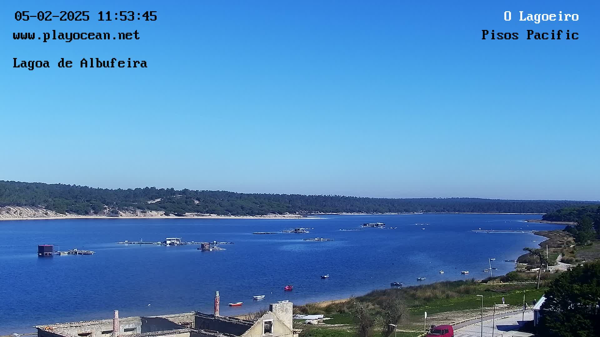 A calm, blue lagoon is seen under a clear blue sky, with several small boats and structures on the water and a sandy shore with sparse vegetation.