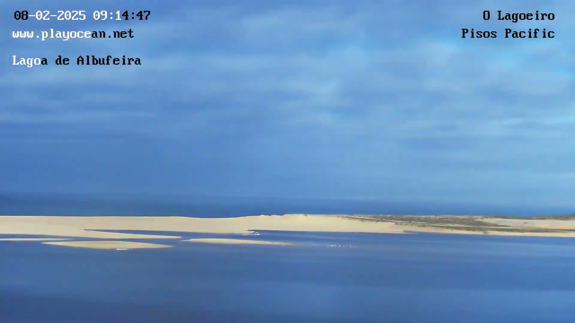 A mostly clear blue sky overlooks a pale sandy shoreline and calm blue water.