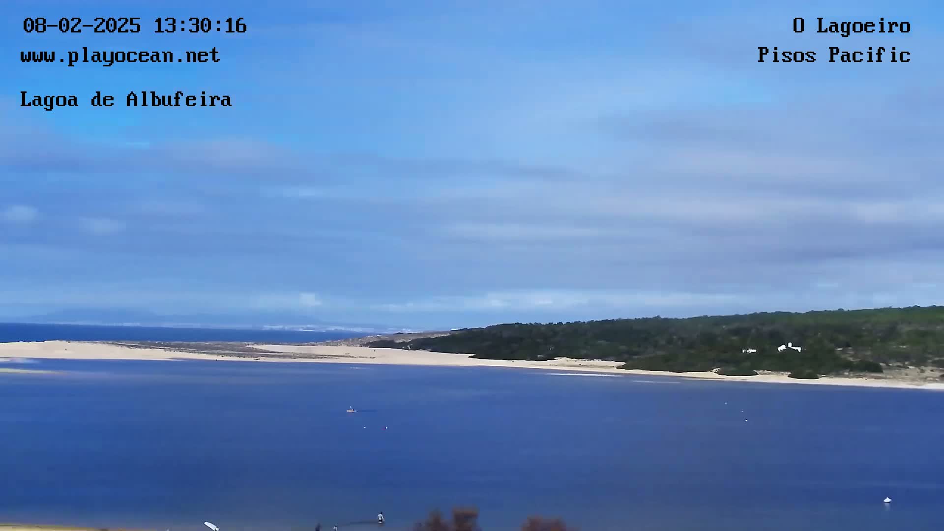 A calm, light-blue lagoon stretches out from a sandy shore lined with green vegetation under a mostly clear blue sky with a few wispy clouds.