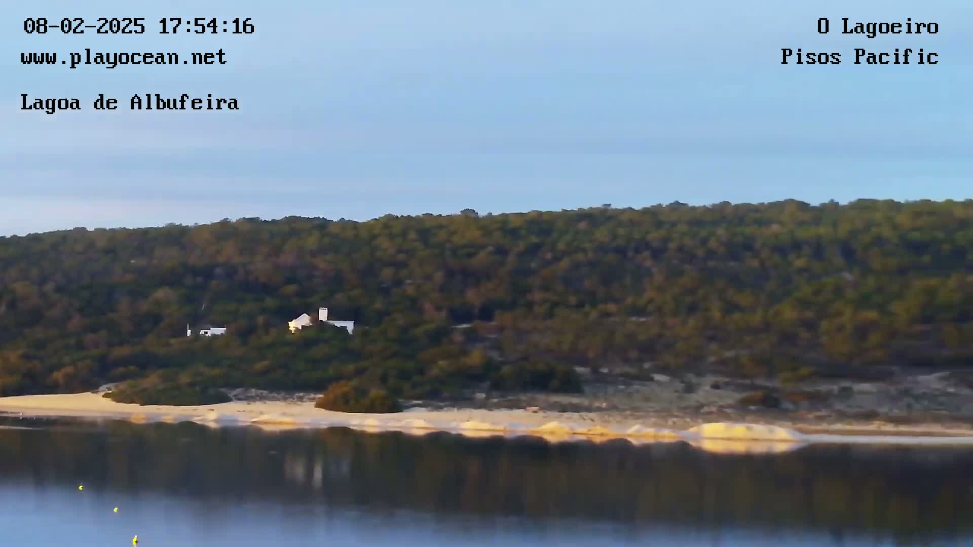A white building sits on a tree-covered hill overlooking a calm body of water with a sandy shoreline under a clear sky.