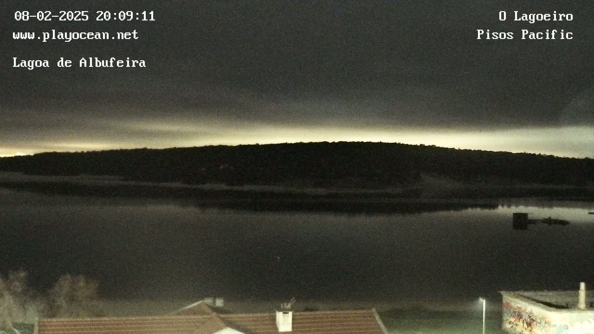 A dark, calm body of water reflects a dimly lit shoreline under a mostly cloudy night sky.