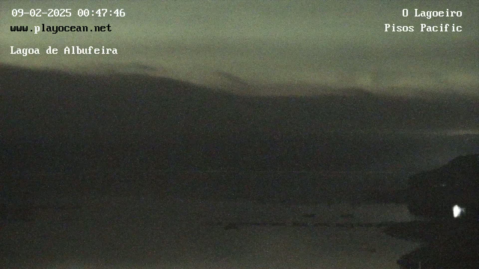 A dark, nighttime view of a body of water, possibly a lagoon, with a distant, dimly lit shoreline under a cloudy sky.