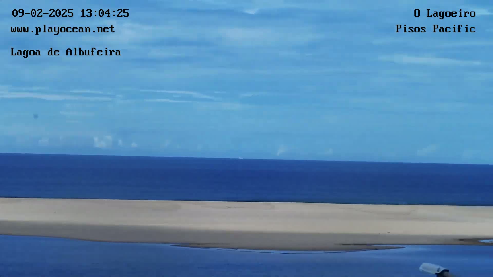 A mostly clear blue sky overlooks a calm blue ocean meeting a long, light beige sandy beach.