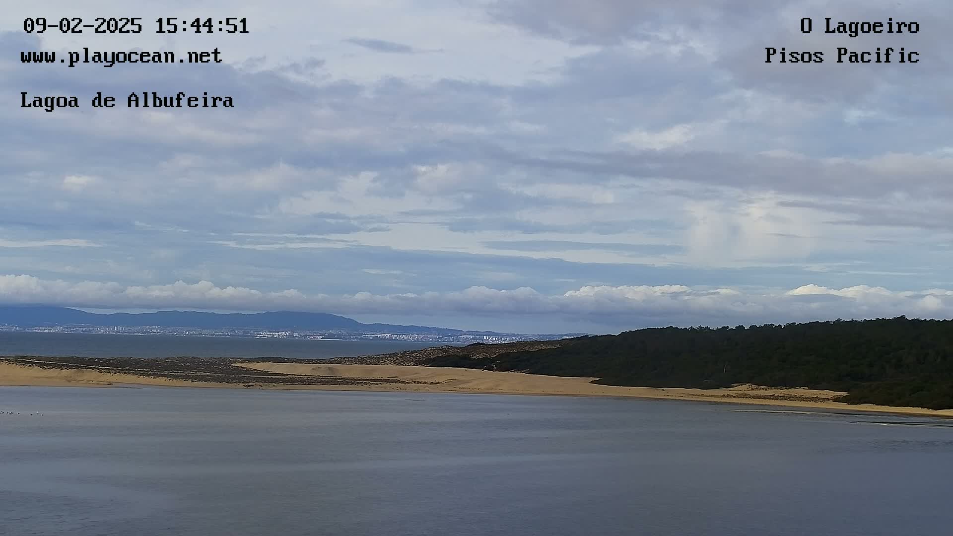 A body of water stretches out before a sandy shore backed by dark green vegetation and distant mountains under a mostly cloudy sky.