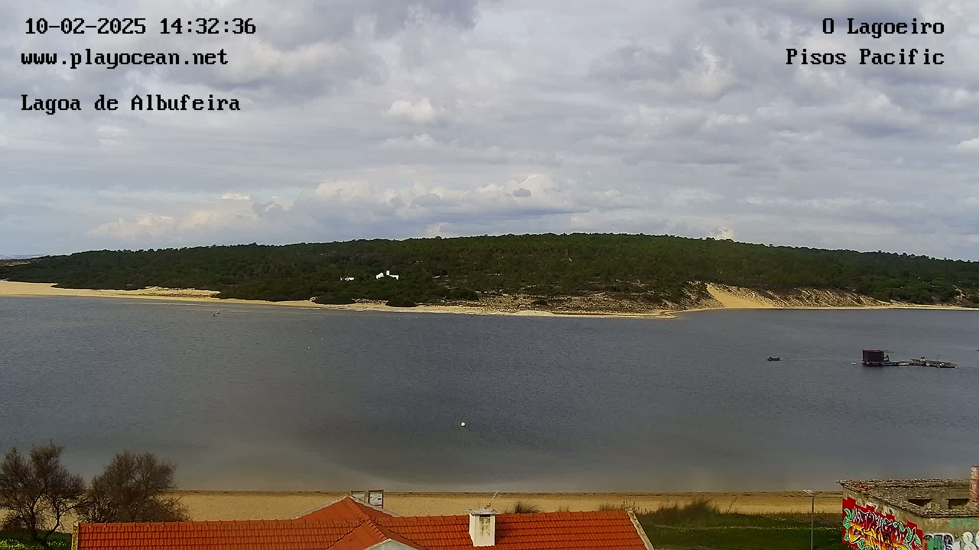 A calm body of water stretches out before a sandy beach and a verdant, tree-covered hillside under a partly cloudy sky.