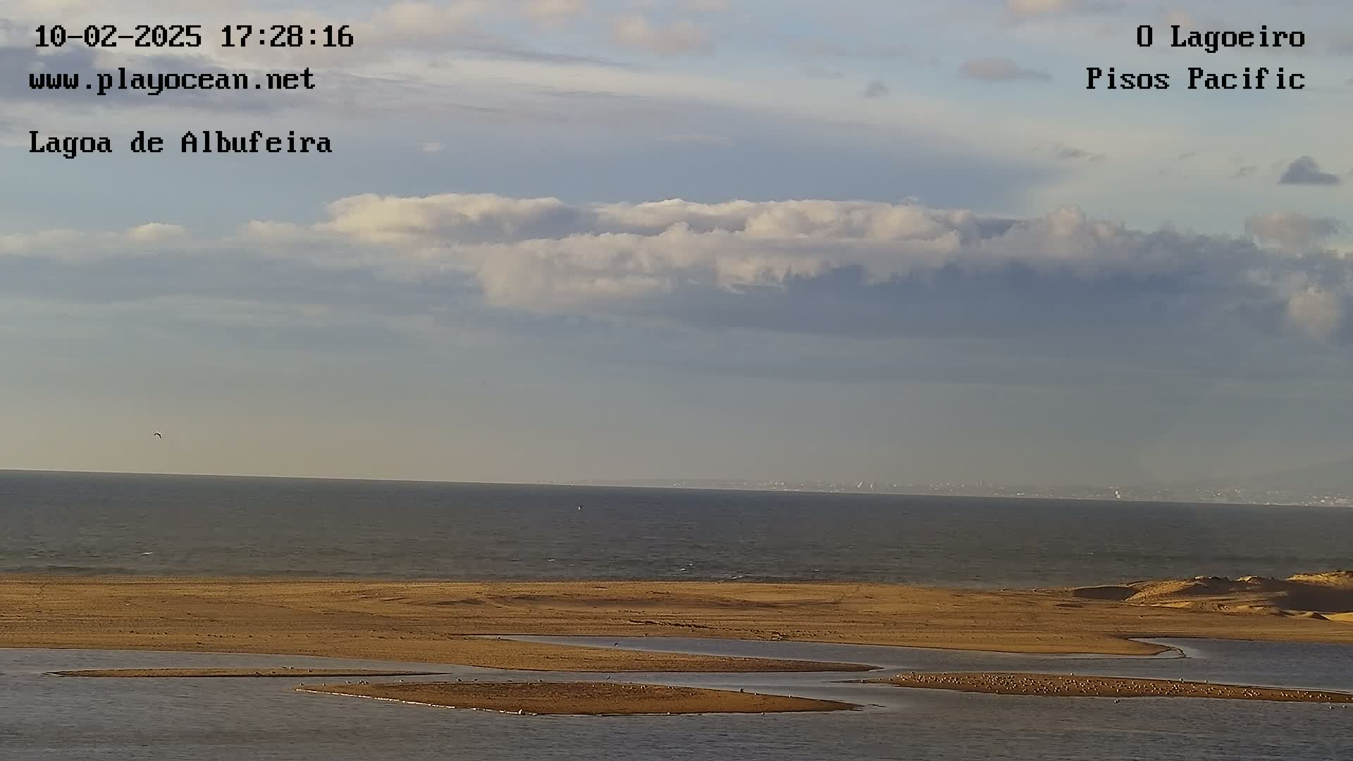 A partly cloudy sky overlooks a calm ocean meeting a sandy shore with several small islands in a lagoon.