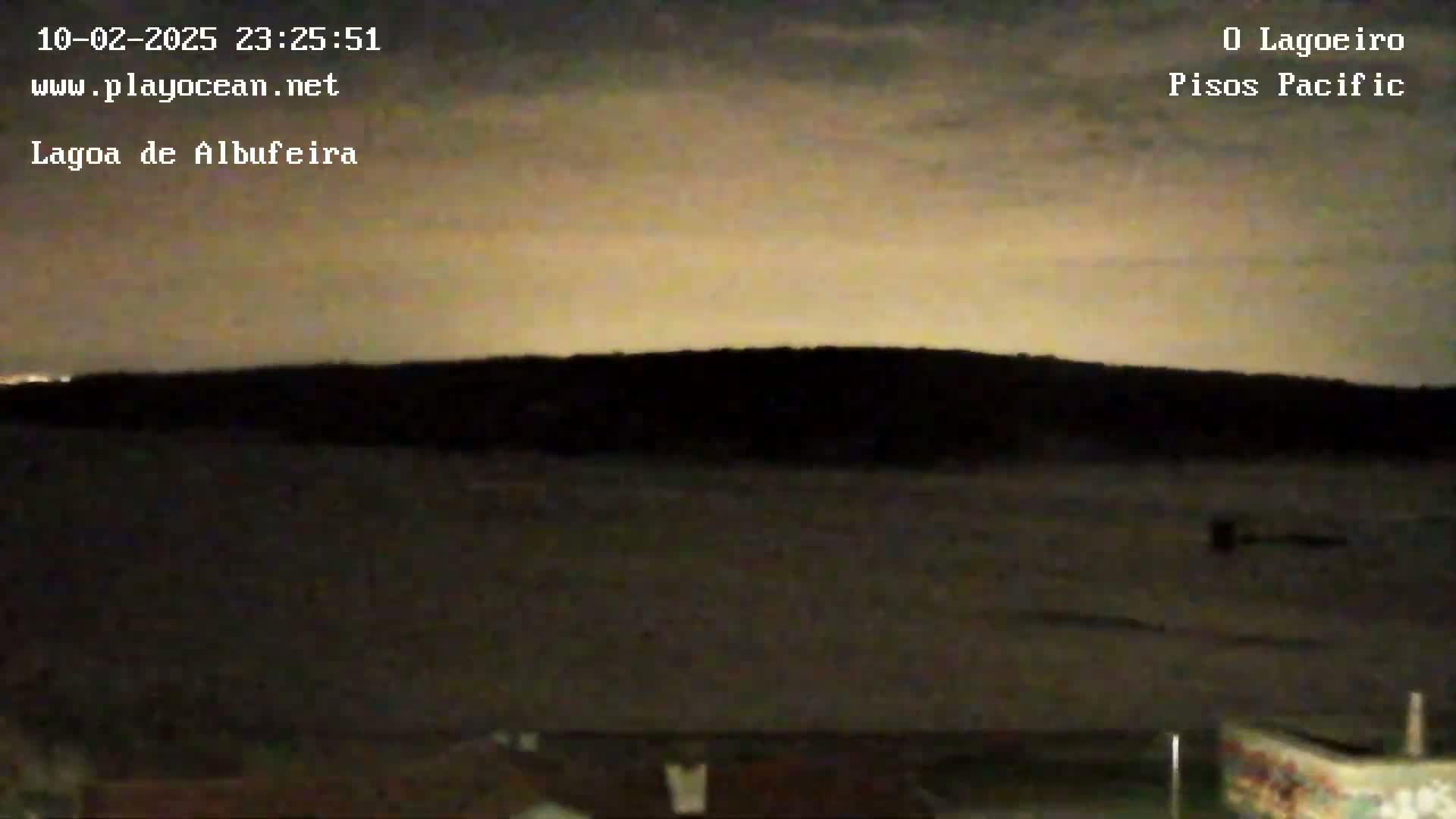 A dark, nighttime landscape shows a body of water in the foreground and a silhouetted shoreline in the background under a mostly cloudy, light-brown sky.