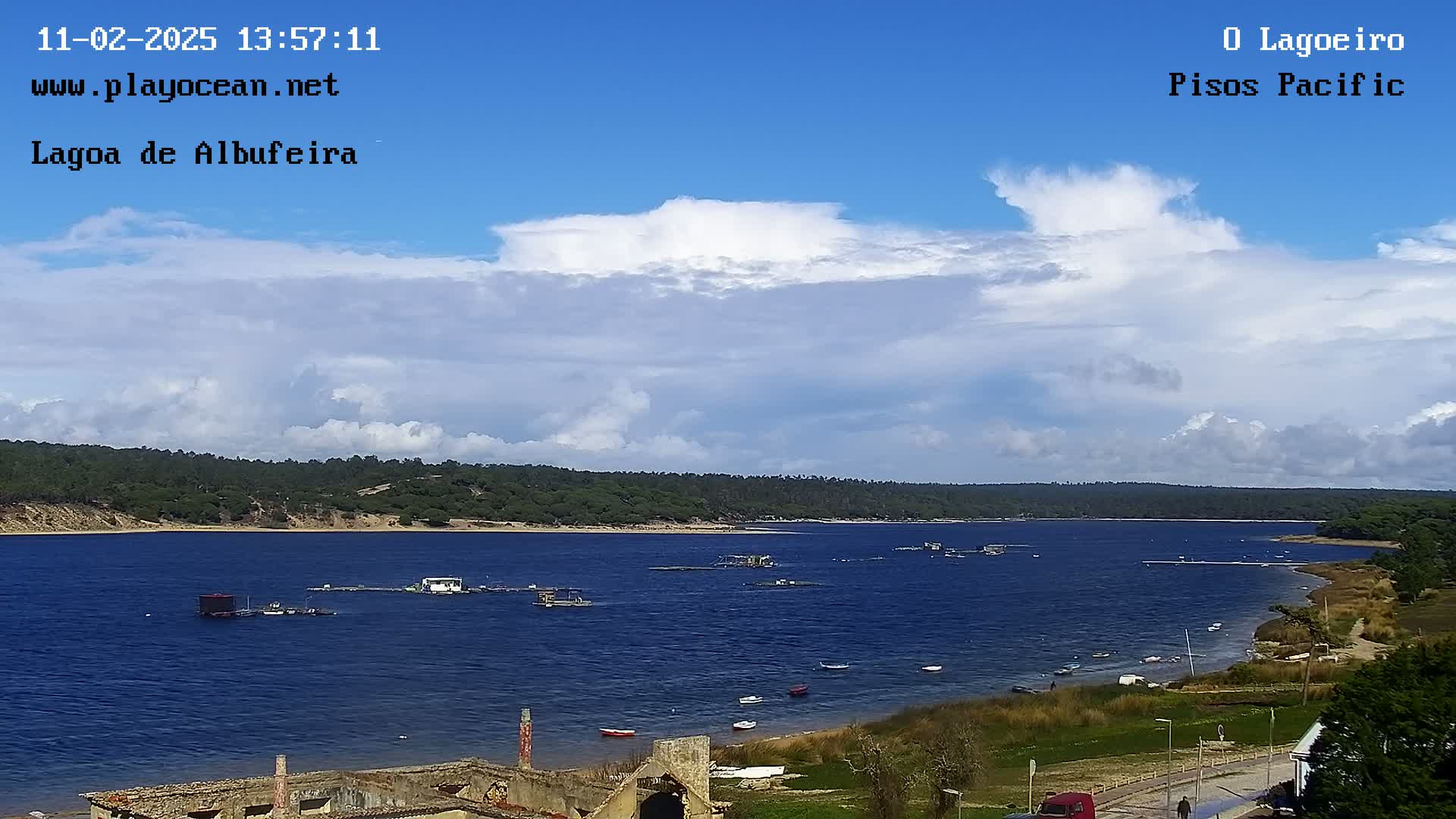 A mostly sunny day with scattered cumulus clouds over a calm body of water with several small boats and structures on the surface, backed by a shoreline of green vegetation.
