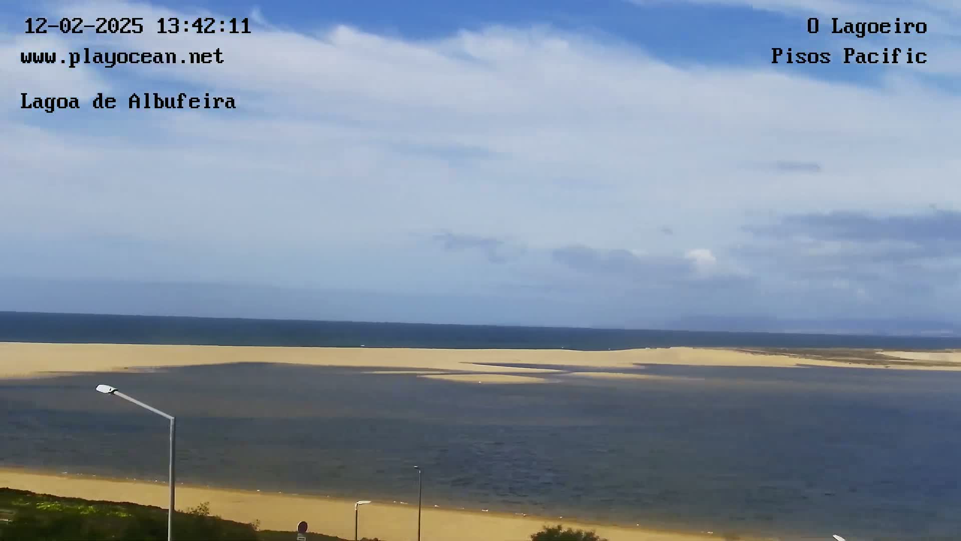 A mostly sunny day shows a wide expanse of calm, dark-blue water separating a sandy beach from a distant ocean horizon.
